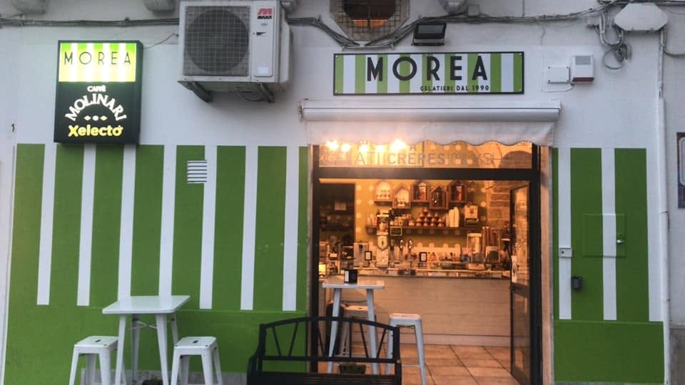 Facciata a strisce verdi e bianche con insegna MOREA Gelatieri dal 1990, porta aperta sul banco illuminato e sgabelli